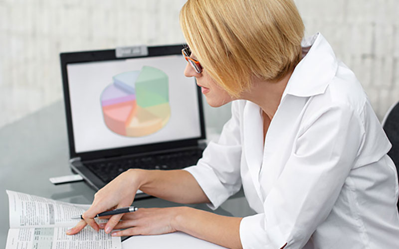 Woman with glasses references book with colorful pie chart on her laptop behind her